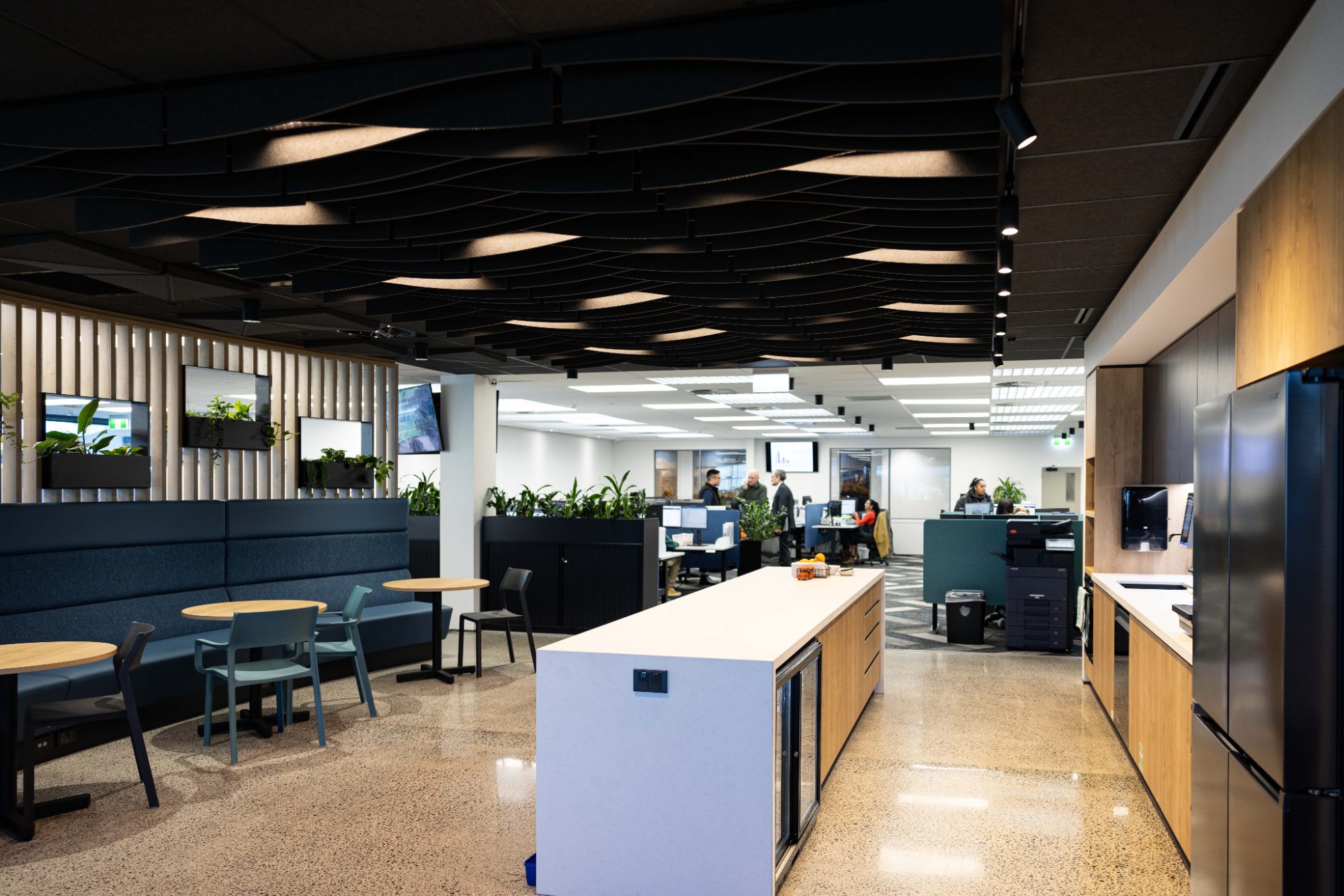 Open-plan office under a dark acoustic ceiling, with a kitchen island and people working at desks