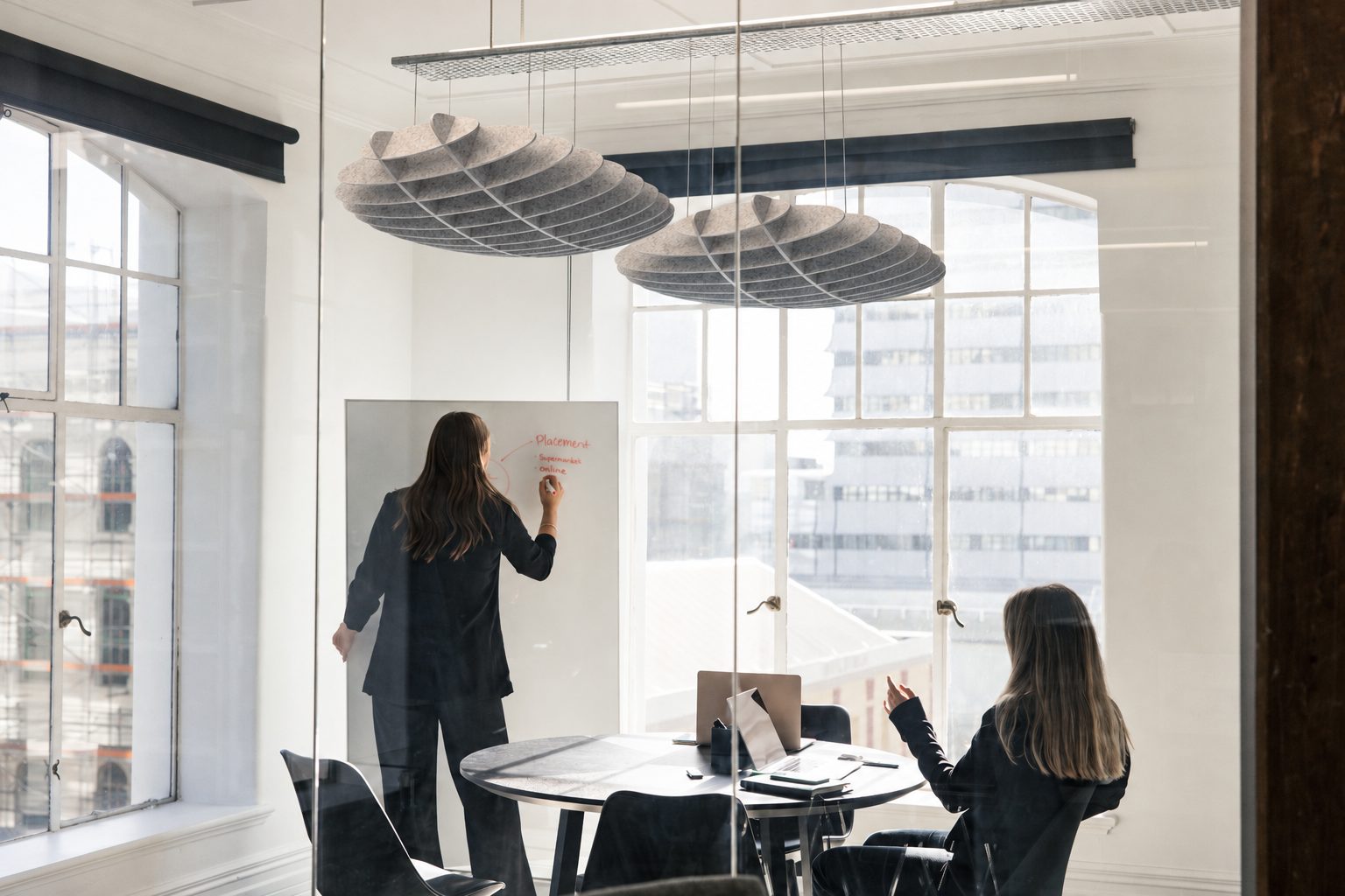 Two women in a glass meeting room under round acoustic pendants, one writing on a whiteboard