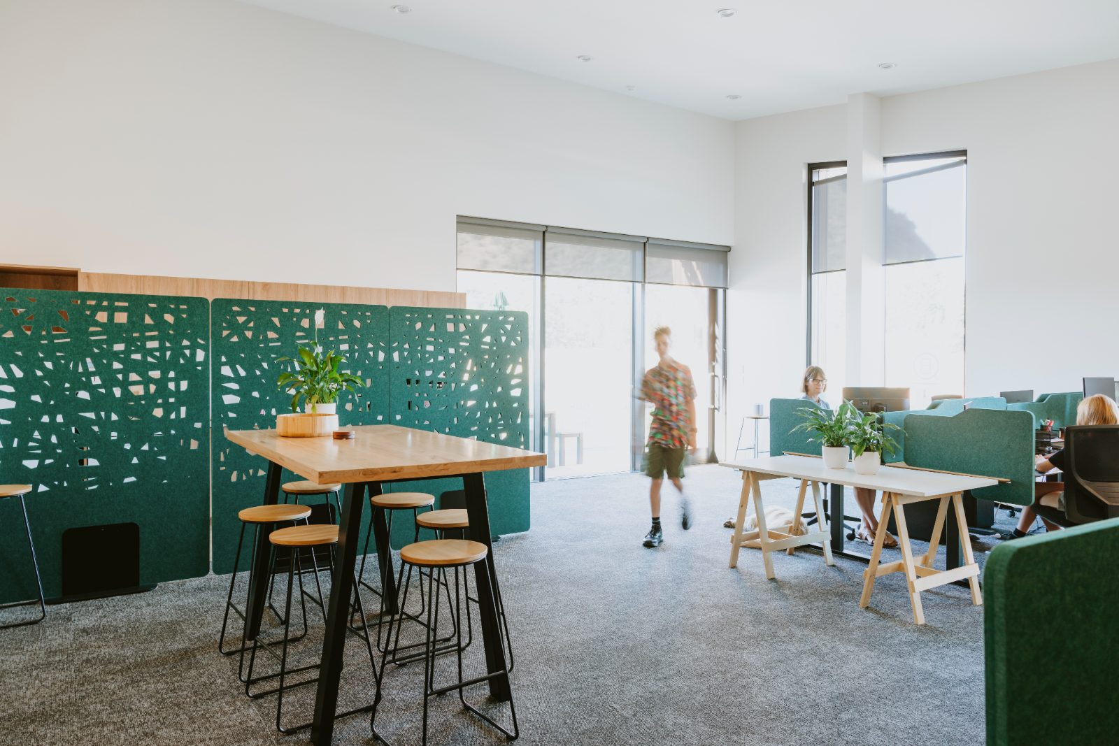 Green acoustic screens with a cut-out pattern in an open office, beside a high table and stools