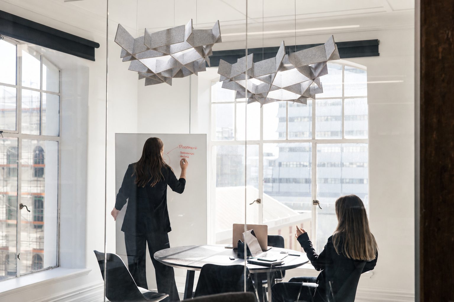 A meeting room with folded grey acoustic pendants over the table, one person writing on a whiteboard