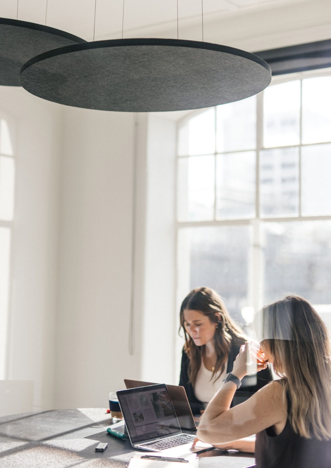 Two people working on laptops at a meeting table under round acoustic pendants